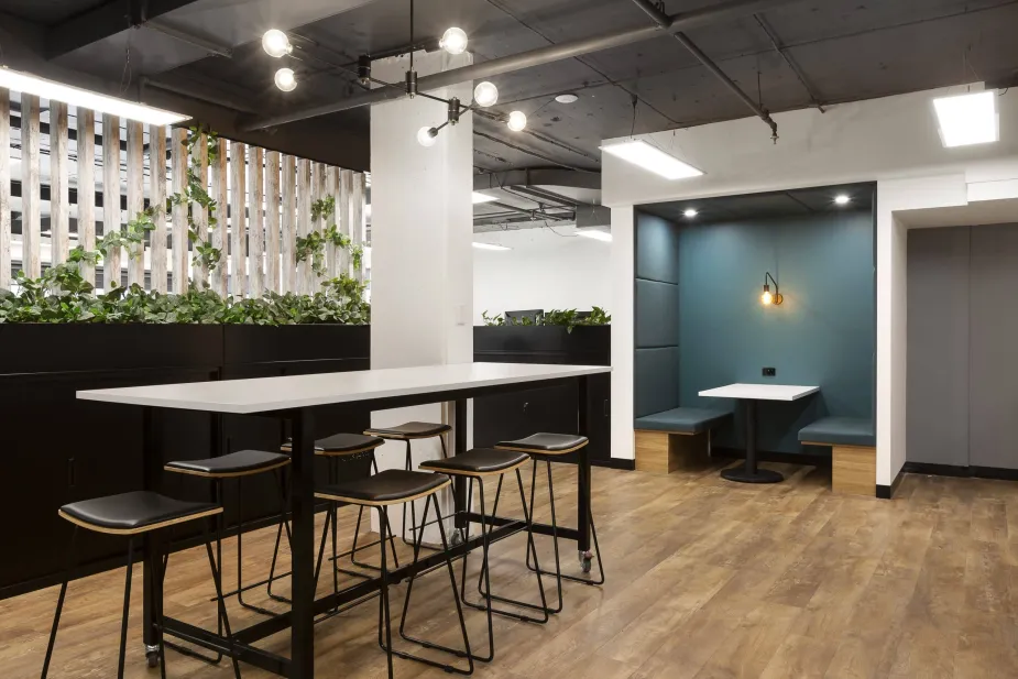 An office break area with high tables, black stools, and a partition wall with green plants, adjacent to a booth seating area.