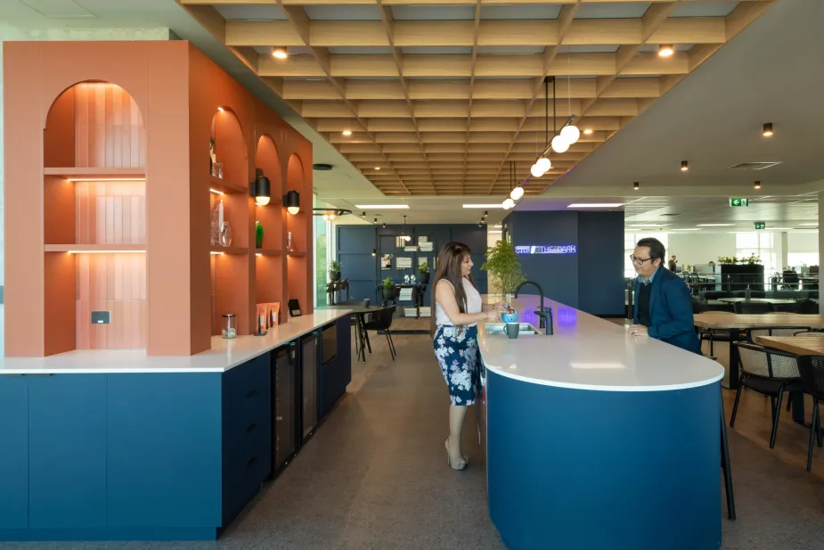 A contemporary office kitchen with a curved blue counter, terracotta shelving units, and modern black bar stools.