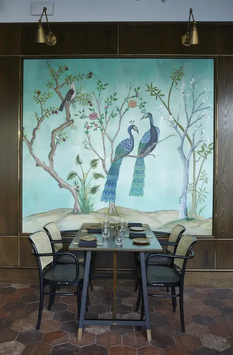 A dining table set against a large, decorative wall painting of two peacocks amidst a floral landscape, with elegant wall-mounted lamps above and terracotta-tiled flooring.