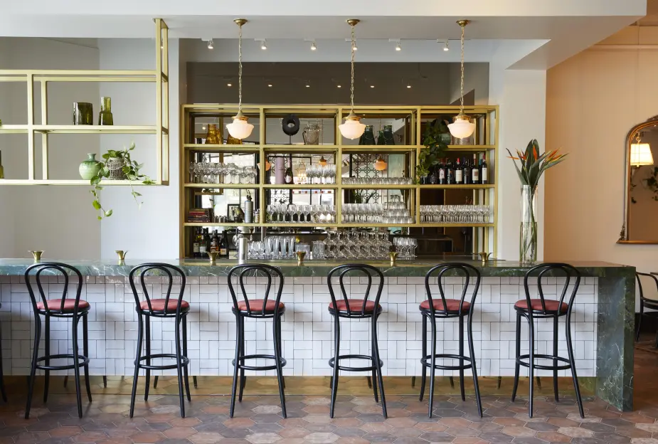A cozy bar area with a tiled counter and high stools, framed by brass shelving with glassware and bottles. Pendant lights hang above, and the space is accented with plants and terracotta floor tiles.