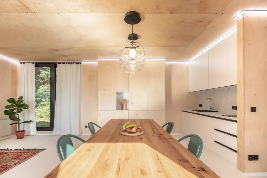 A bright, cozy interior with a natural wood dining table, teal chairs, and a kitchen with white cabinetry against a backdrop of plywood walls and ceiling.