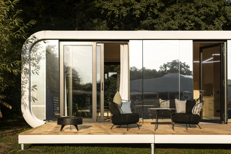 An exterior view of a modern modular home with curved corners, expansive glass windows, and a sliding door that opens onto a wooden deck with a fire pit and two metal frame chairs with cushions