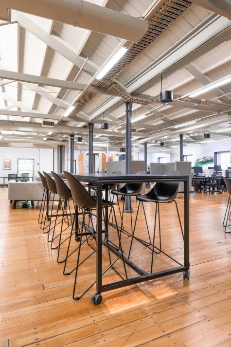 A spacious modern office with an open floor plan, featuring wooden flooring, black desks with computer setups, and exposed ceiling beams.