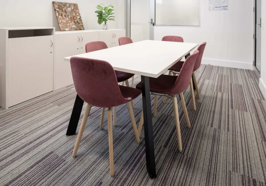 A modern meeting room featuring a white rectangular table with black legs, surrounded by four purple chairs with wooden legs