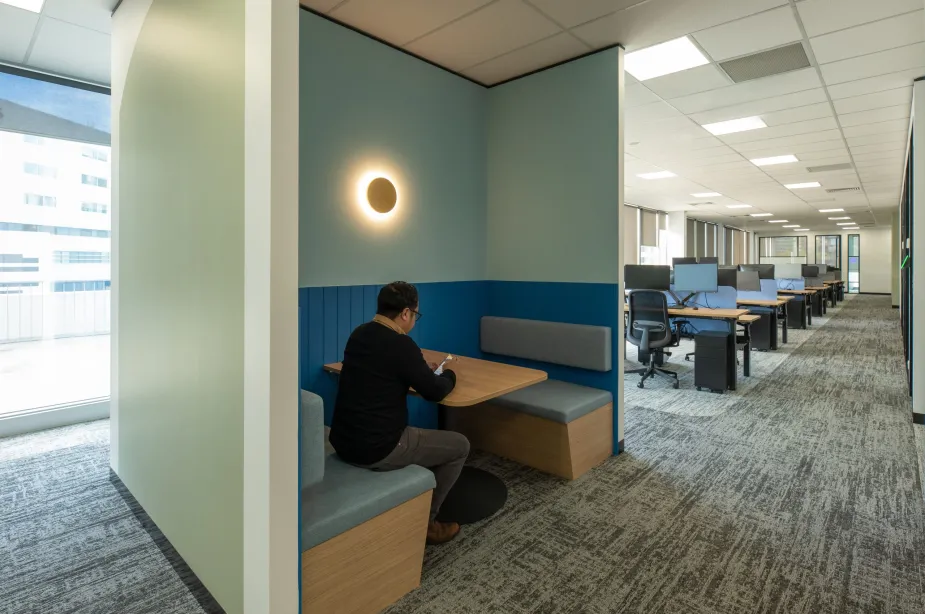 An office breakout area featuring a booth-style seating with a staff sitting at a wooden table, focused on reading or writing.