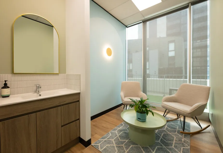 An office relaxation area with two comfortable chairs, a round coffee table with a plant, a mirror on the wall, and a view of the city through large windows.