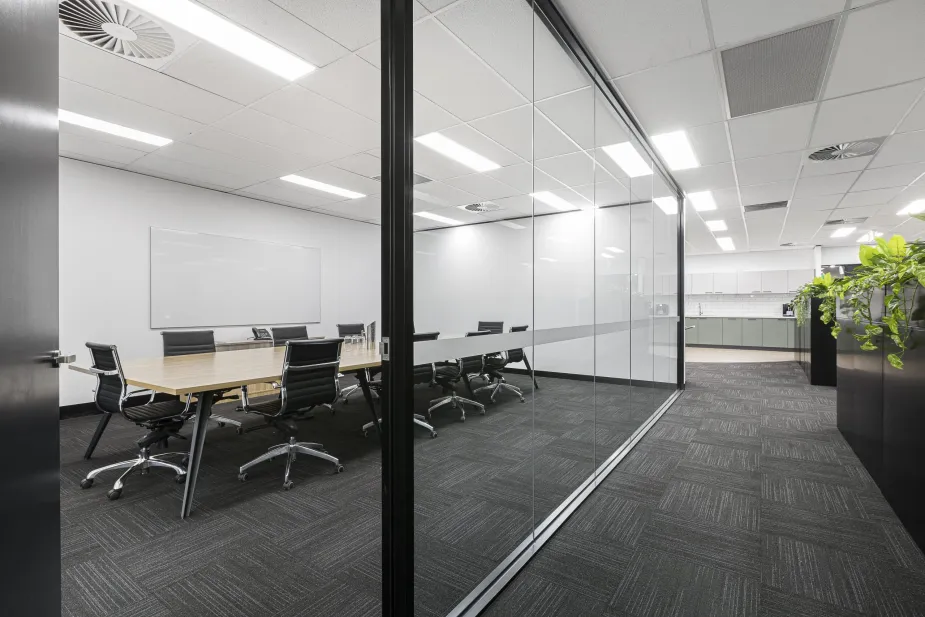 A view into a modern conference room through glass walls, showing a long table with chairs and a whiteboard on the wall.