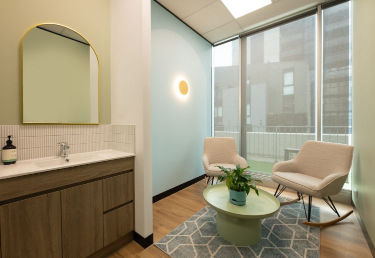 An office relaxation area with two comfortable chairs, a round coffee table with a plant, a mirror on the wall, and a view of the city through large windows.
