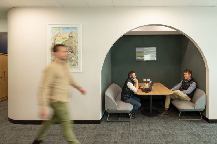 Two people having a discussion in a semi-private office nook with a man walking past in the foreground.