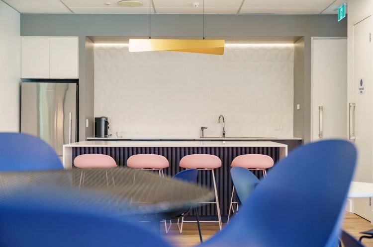 Office kitchenette with pink stools under a counter, a yellow overhead light, and a fridge to the side
