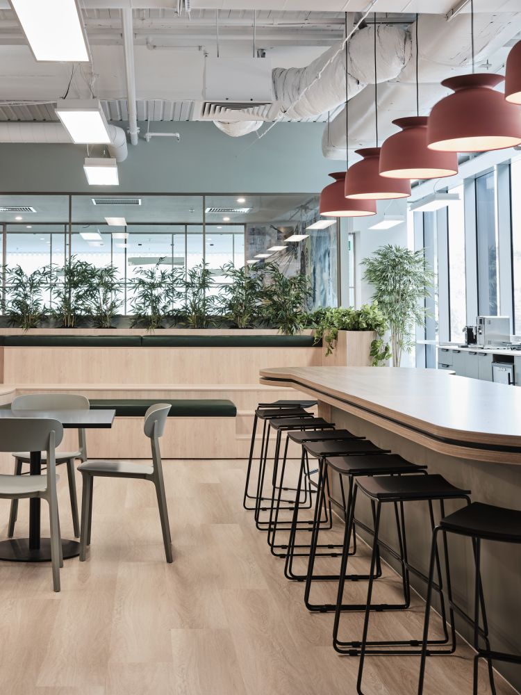 A modern office cafeteria with hanging red lamps, bar stools, and greenery.
