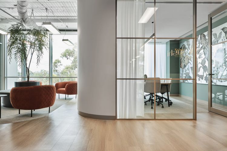 An office lounge with red armchairs and large windows, adjacent to a meeting room with transparent glass walls and nature-inspired art.
