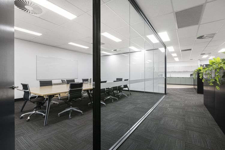 A view into a modern conference room through glass walls, showing a long table with chairs and a whiteboard on the wall.
